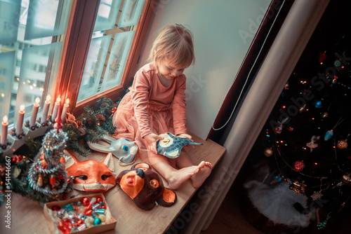Cute little girl is dressing up a tiny Christmas tree and trying on New Year's animal masks. Image with selective focus and toning. 