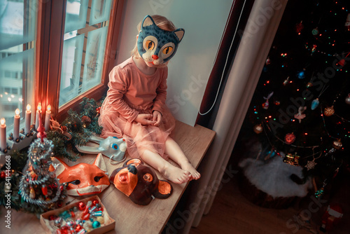 Cute little girl is dressing up a tiny Christmas tree and trying on New Year's animal masks. Image with selective focus and toning. 