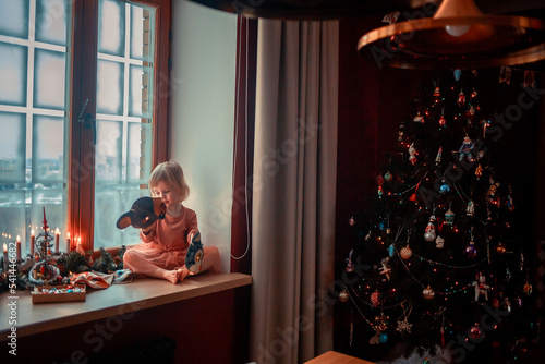 Cute little girl is dressing up a tiny Christmas tree and trying on New Year's animal masks. Image with selective focus and toning. 