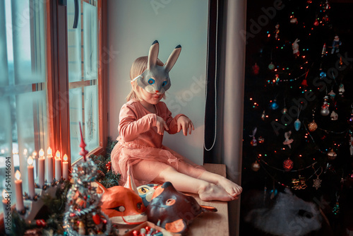 Cute little girl is dressing up a tiny Christmas tree and trying on New Year's animal masks. Image with selective focus and toning. 