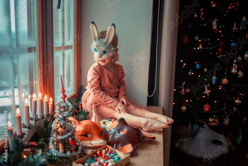 Cute little girl is dressing up a tiny Christmas tree and trying on New Year's animal masks. Image with selective focus and toning. 