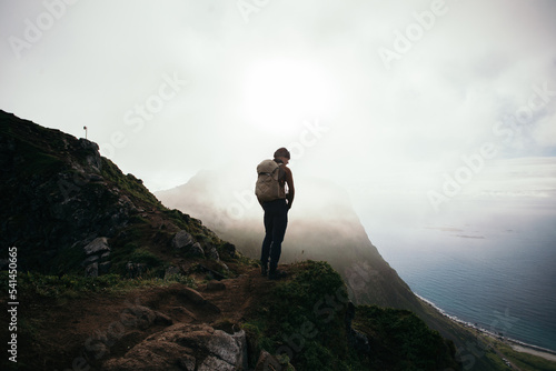 Wallpaper Mural Hiker stand on top of mountain climb. Man in hiking outfit with backpack stand on top of hill overlooking epic mountain views covered by clouds. Hiking in Norway, Lofoten Torontodigital.ca