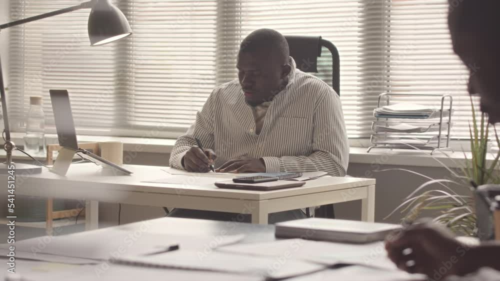 Serious African American businessman filling in financial documents sitting at desk in modern office Serious African American businessman filling in financial documents sitting at desk in modern office