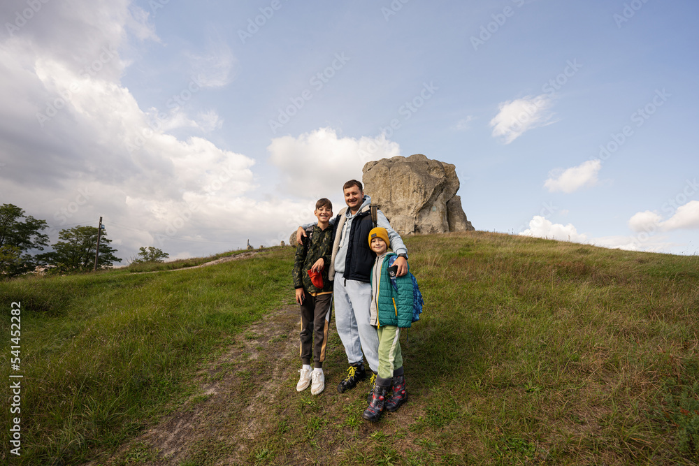 Father with two sons wear backpack hiking near big stone in hill. Pidkamin, Ukraine.