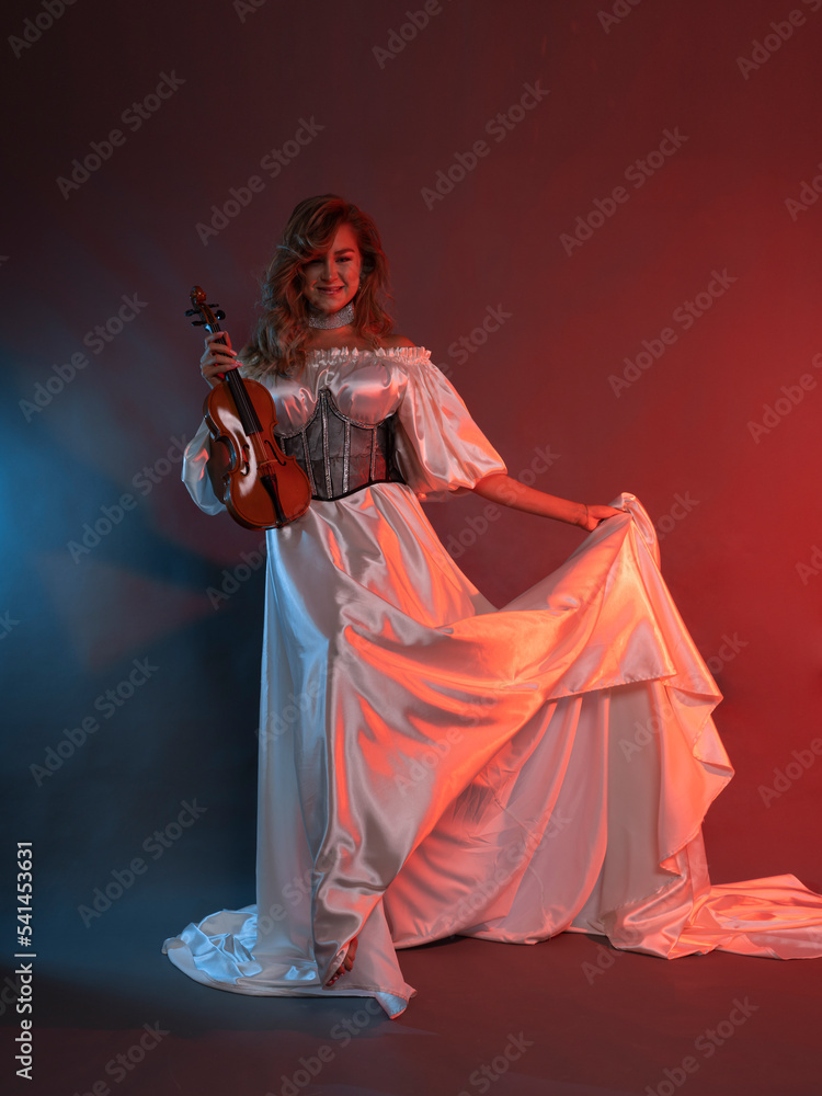 Fototapeta premium A woman poses in a long white silk dress on a colored background with a violin in her hands. A model in a flowing dress. Portrait of a full-length beauty.