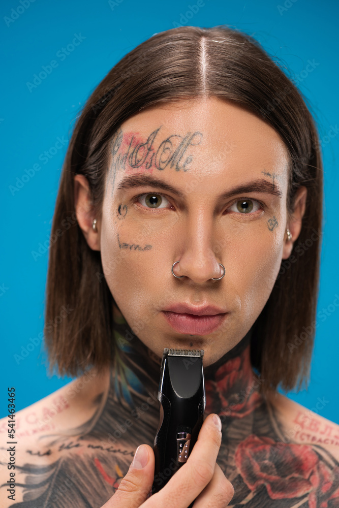 young and pierced man with tattoos trimming face with electric razor isolated on blue young and pierced man with tattoos trimming face with electric razor isolated on blue