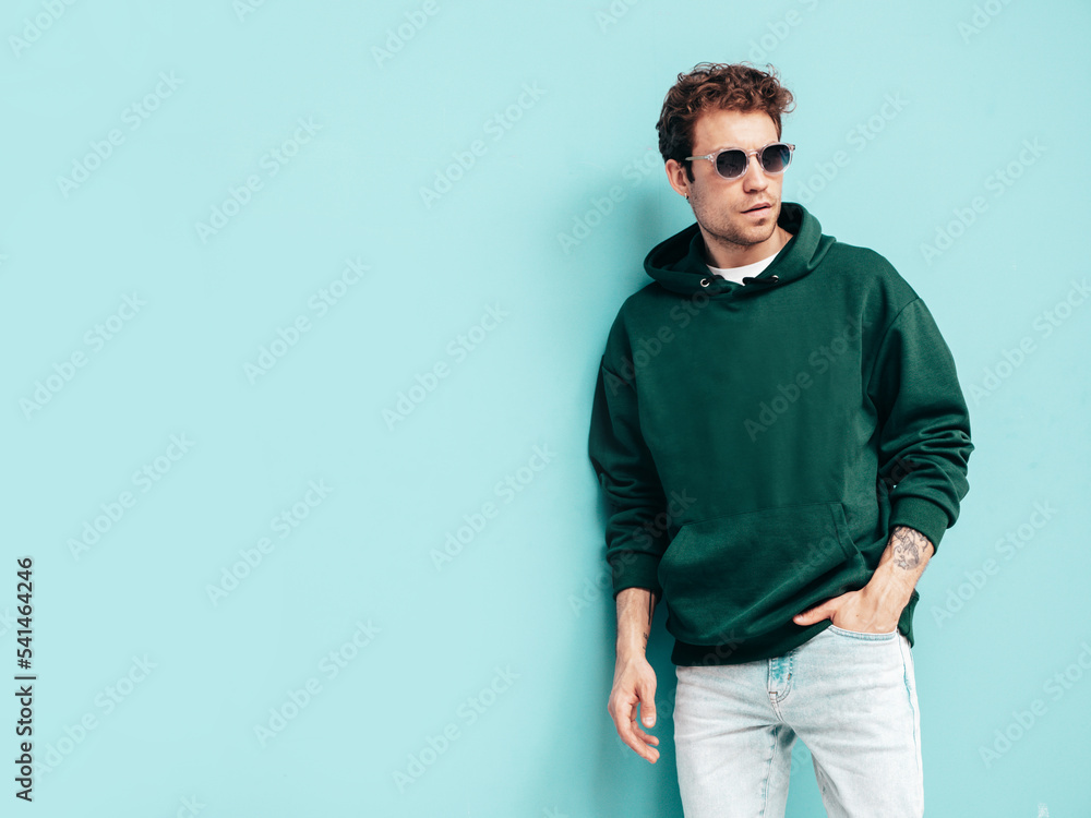 Handsome confident hipster model.Sexy unshaven man dressed in summer stylish green hoodie and jeans clothes. Fashion male with curly hairstyle posing in studio. Isolated on blue. In sunglasses Handsome confident hipster model.Sexy unshaven man dressed in summer stylish green hoodie and jeans clothes. Fashion male with curly hairstyle posing in studio. Isolated on blue. In sunglasses