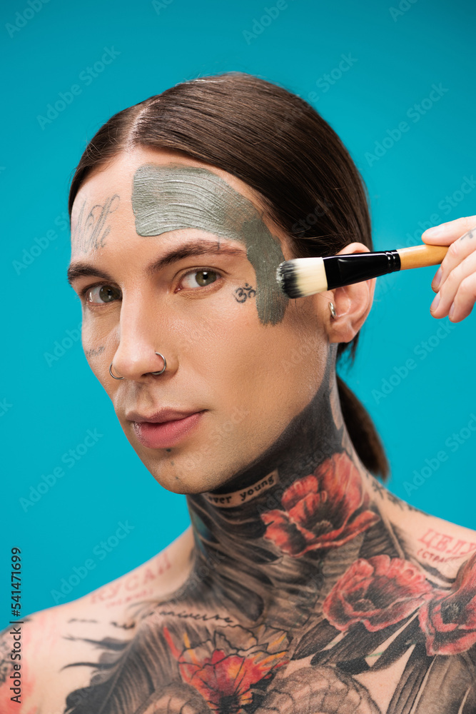 Obraz premium pierced young man with tattoos applying clay mask with cosmetic brush isolated on blue
