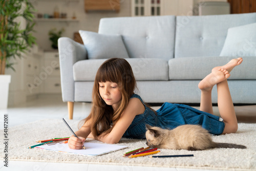 A cute funny girl in a denim sundress and bangs in her hair lies in a room on a carpet and draws with colored pencils. Next to her is a four-legged friend - a small playful fluffy kitten