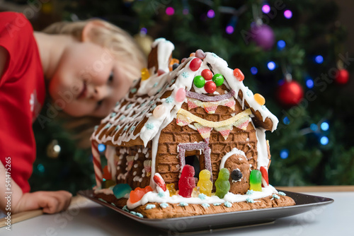 Toddler looking at Homemade gingerbread house decorated with icing, sweets , and jelly candies with blurred Christmas tree on background.