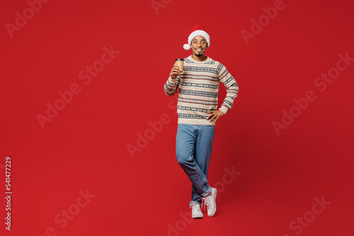 Konstfotografi Full body merry young man wear Christmas sweater Santa hat posing hold takeaway delivery craft paper cup coffee to go isolated on plain red background