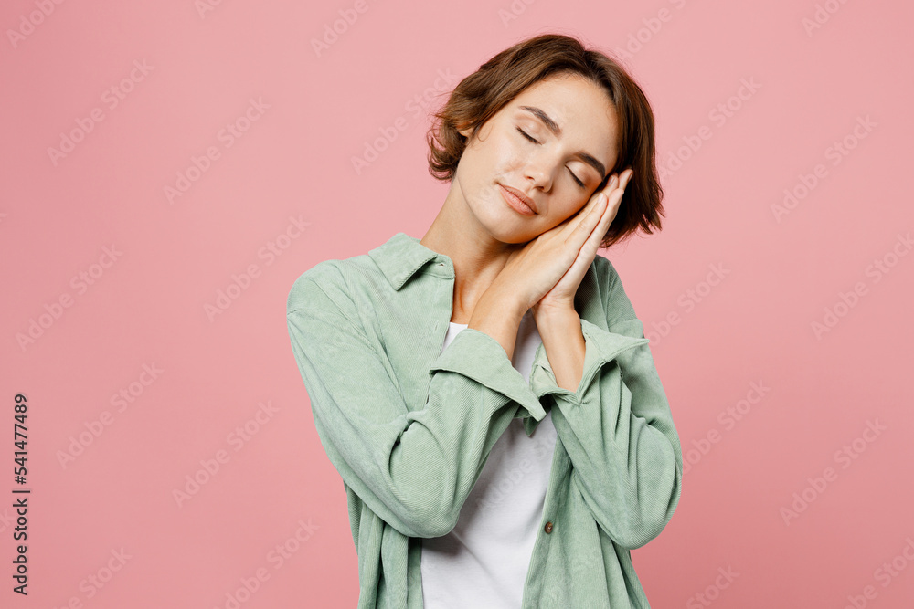 Young calm sleepy woman 20s she wearing green shirt white t-shirt close eyes holding folded hands under cheek isolated on plain pastel light pink background studio portrait. People lifestyle concept. Young calm sleepy woman 20s she wearing green shirt white t-shirt close eyes holding folded hands under cheek isolated on plain pastel light pink background studio portrait. People lifestyle concept.