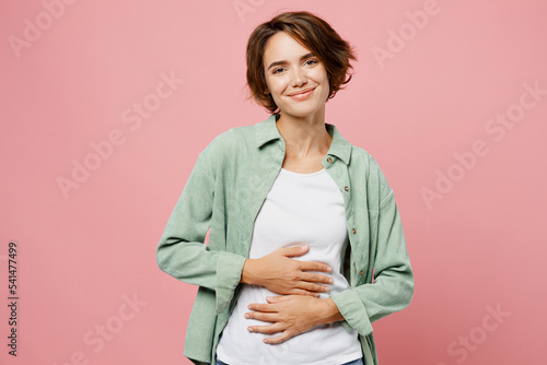 Young smiling cheerful fun happy woman 20s she wear green shirt white t-shirt holds hands on belly look camera isolated on plain pastel light pink background studio portrait. People lifestyle concept. photo