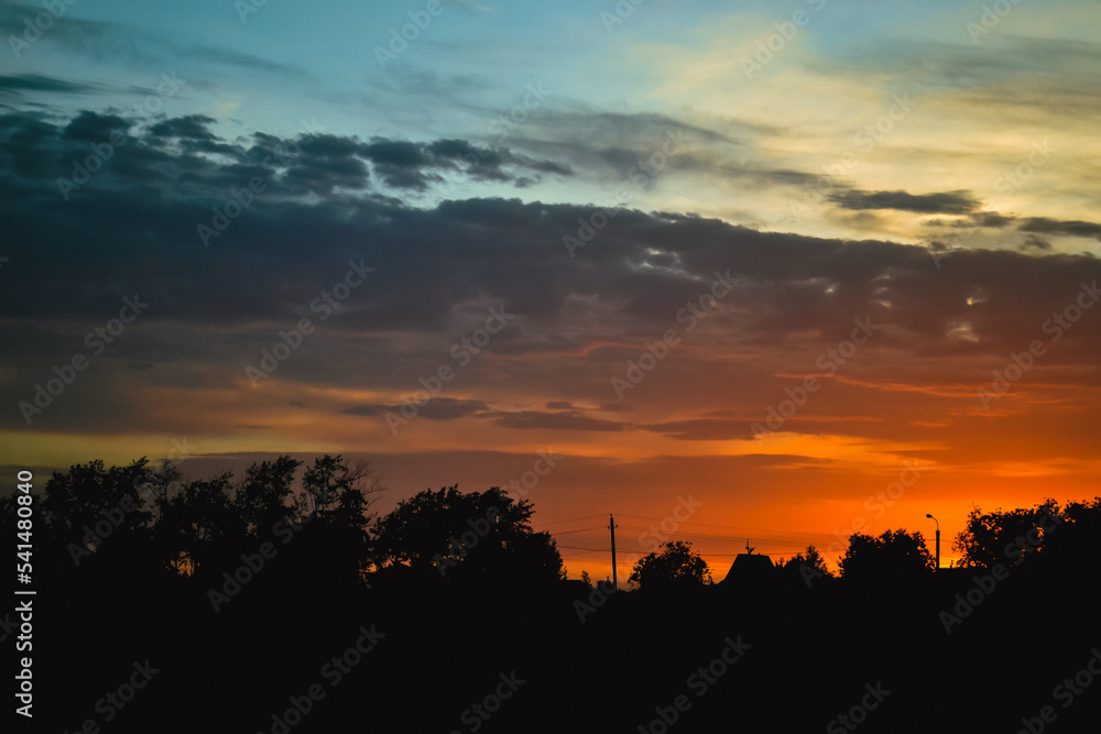 Morning glow illuminates the clouds on a beautiful colorful sky above the silhouettes of trees, a stob and a triangular roof of a house at dawn