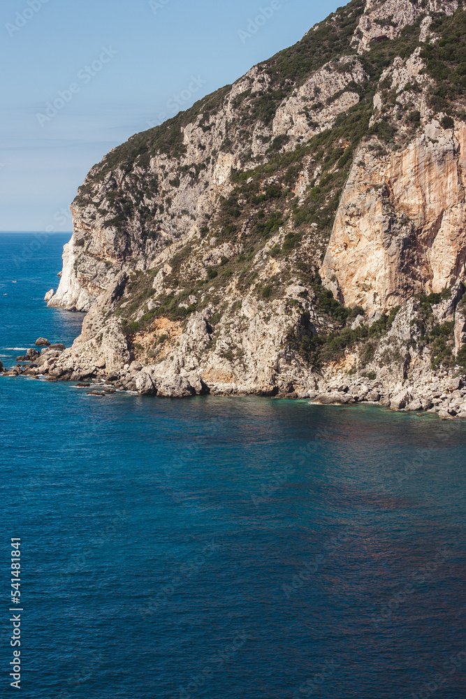 Corfu, Greece. A huge mountain rock on which green grows grows near beautiful blue water where the summer sun shines.