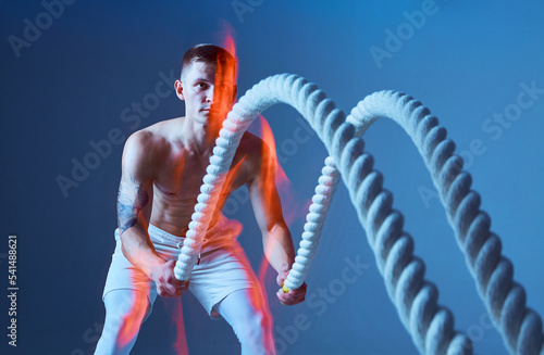 Young muscular half naked sportsman pull the rope on blue background. Rope on the foreground.