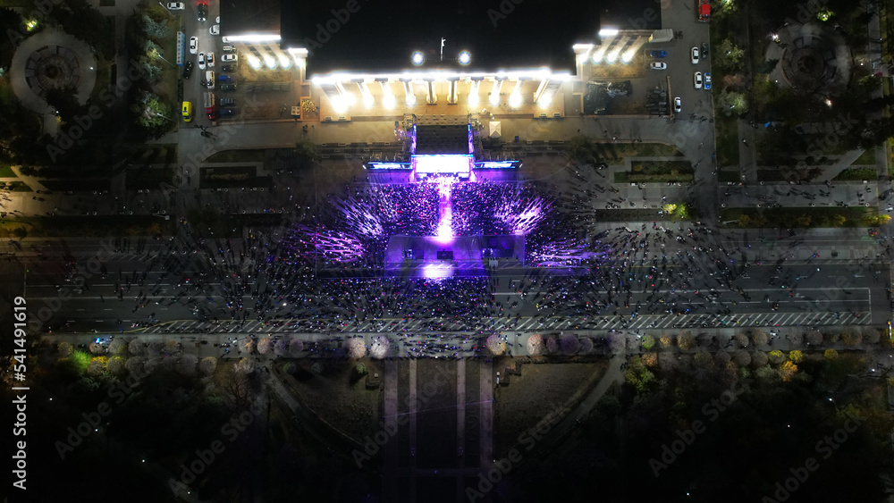 Top view from a drone of a big concert in the night city. Lots of people, rays of light. A large building with lighting. There are streets with cars, pedestrian streets with people and a park. Almaty Top view from a drone of a big concert in the night city. Lots of people, rays of light. A large building with lighting. There are streets with cars, pedestrian streets with people and a park. Almaty