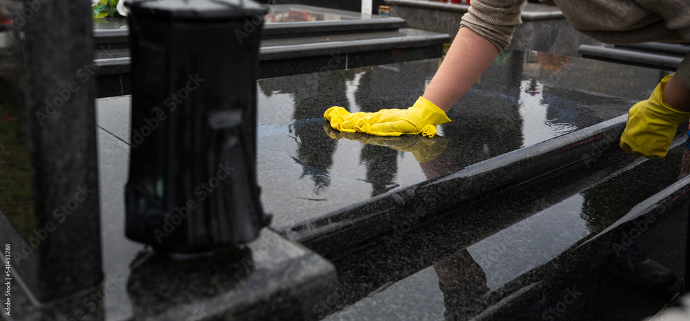 Headstone cleaning on cemetery. Professional in yellow gloves cleans the marble grave, polishing marble tombstone with special liquid, preparation for All Saints Day on November 1st