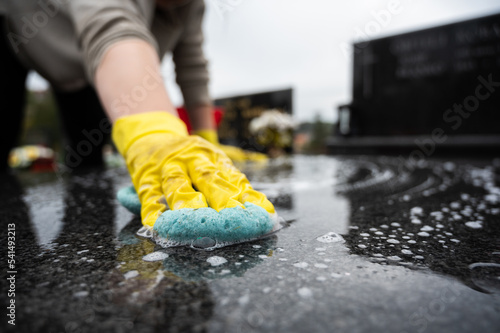 Headstone cleaning on cemetery. Professional in yellow gloves cleans the marble grave, scrubbing with sponge and water. Tombstone preparation for All Saints Day on November 1st