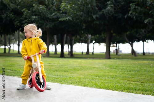 Happy active pretty cute caucasian blonde baby girl, kid, toddler,smiling child about 2 years old wearing bright yellow jumpers learning riding run balance bike in summer park outdoors.