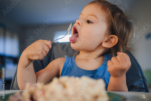 Baby girl sitting at the children s table  eating with appetite and licking the spoon. Healthy eating. Child health care. Sweet food.