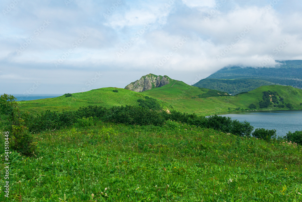 Obraz premium natural landscape of Kunashir island with grassy hills, volcanic rocks, volcano in the clouds, lagoon and ocean in the distance
