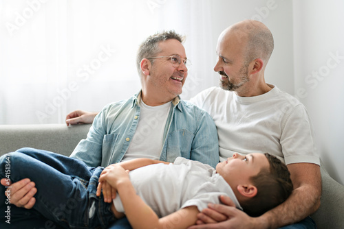 Happy two man couple with adopted child sit on sofa at home