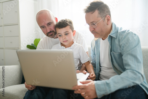 Happy two man couple with adopted child sit on sofa at home using laptop