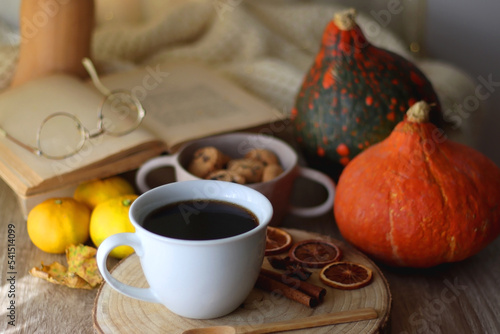 Cup of tea or coffee, seasonal spices, bowl of cookies, blanket, pumpkins, colorful leaves, books and tangerines on wooden table. Cozy hygge at home. Selective focus.