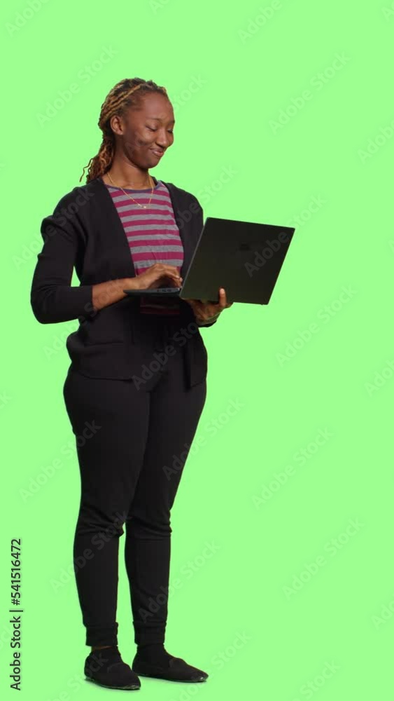 Vertical video: African american girl using laptop and browsing internet, holding wireless pc to scroll through online website. Smiling female model standing over full body greenscreen backdrop. Vertical video: African american girl using laptop and browsing internet, holding wireless pc to scroll through online website. Smiling female model standing over full body greenscreen backdrop.