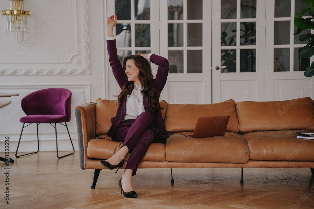 Obraz premium Amazed beautiful brunette woman in violet suit spread her hands in surprise expression with opened mouth, sitting on sofa at home, remote video call, task completed, successful manager celebrating.