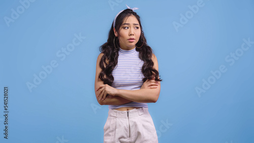 worried asian woman in headband with bow standing with crossed arms isolated on blue