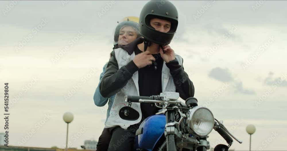 A young stylish couple of bikers sit on a bike standing in a public city parking. The guy and the girl put on motorcycle helmets. Love and safety A young stylish couple of bikers sit on a bike standing in a public city parking. The guy and the girl put on motorcycle helmets. Love and safety