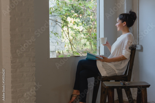 Beautiful young Asian woman drinking coffee and looking through window while sitting on a chair near window at home.