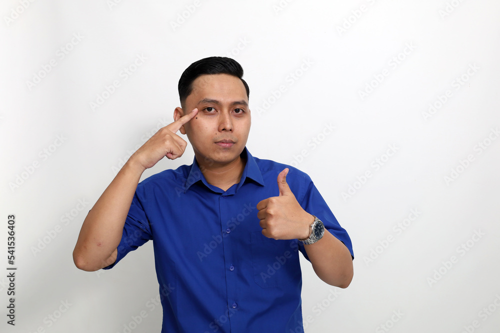 Happy young asian man standing while pointing his eyes and giving thumbs up. Isolated on white background Happy young asian man standing while pointing his eyes and giving thumbs up. Isolated on white background