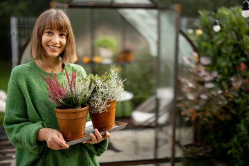 Portrait of young cheerful woman holding flower pots with herbs while standing in front of glasshouse at backyard. Hobby, growing plants concept