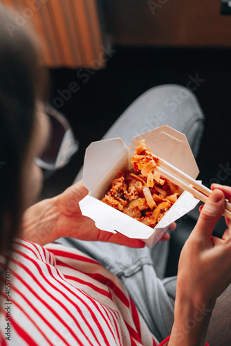 Woman eating fast food from takeaway box asian chinese noodles with vegetables in wok box using chopsticks. Contactless Food delivery. Girl Enjoying Take away udon meal lunch in home apartment.