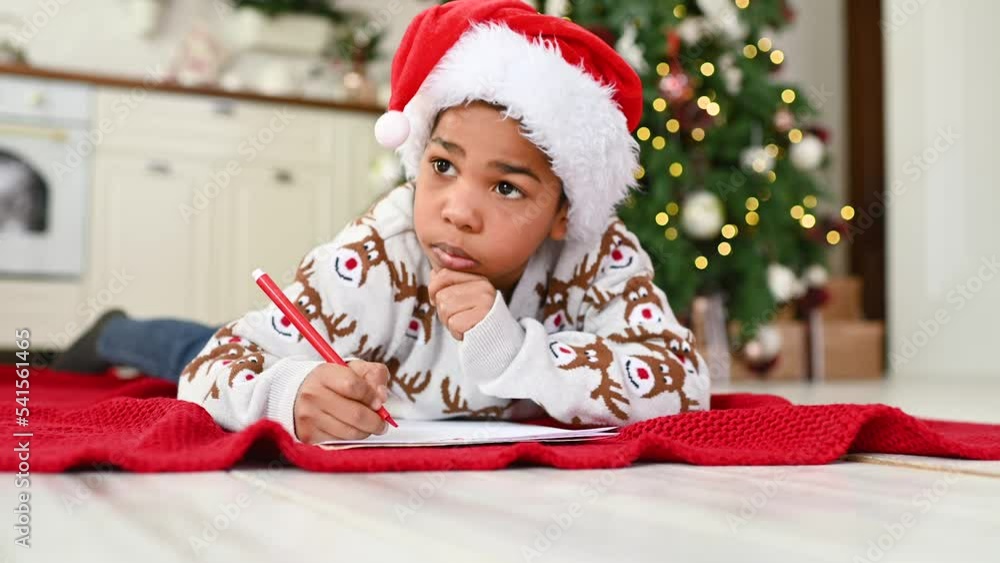 child boy in a santa hat lying on a plaid smiles dreamily and writes a letter to Santa on a white sheet - a wish list on the background of a Christmas tree. child boy in a santa hat lying on a plaid smiles dreamily and writes a letter to Santa on a white sheet - a wish list on the background of a Christmas tree.