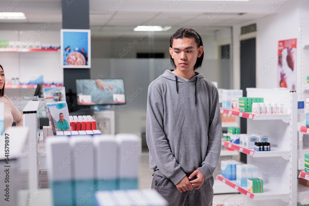 Asian young man looking at medicine boxes on pharmacy shelves, examining supplements to buy prescription treatment. Analyzing medical supplies and medicaments products at health drugstore.