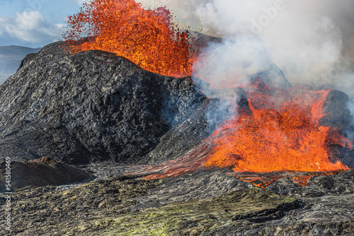 Direct view of the spouting volcano with lava fountains on Iceland. Active volcano in the day. Volcano erupting with strong lava flow. Dark rock around the crater. Steam from the volcano crater