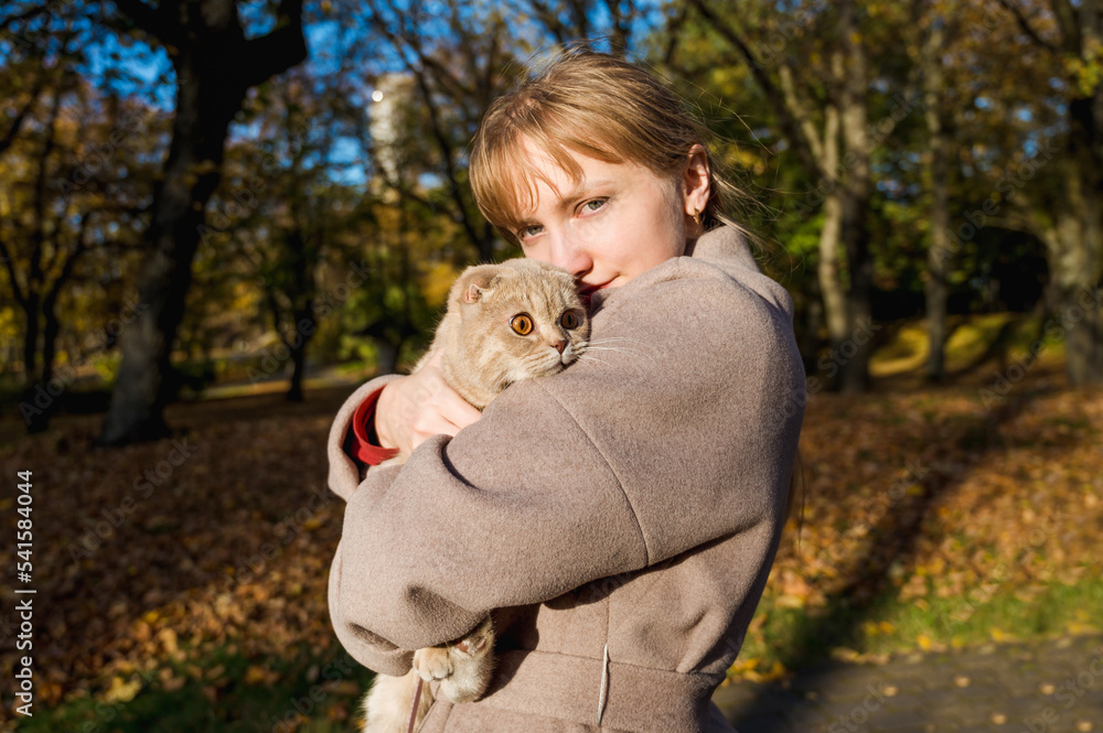 Young woman in beige coat holds a cream-colored Scottish Fold cat in her arms in autumn park at sunny day. Beautiful pet. Domestic cat. Young woman in beige coat holds a cream-colored Scottish Fold cat in her arms in autumn park at sunny day. Beautiful pet. Domestic cat.