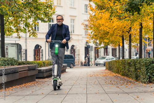 Middle-aged gray-haired man in formal clothes riding rental electric scooter to work through city center with trees around. Environmentaly conscious. Horizontal outdoor shot. High quality photo