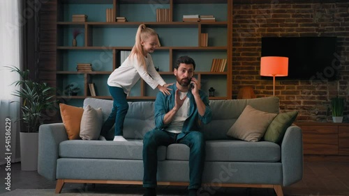 Irritated angry businessman freelancer tired father talk phone freelance work at home during coronavirus lockdown restless little noisy kid girl child need attention disturb man jumping to distracting photo