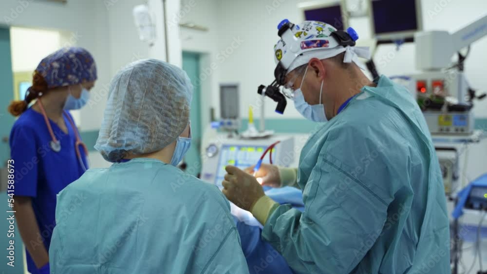 Medics performing surgery standing their backs to camera. Surgeon inserts the instrument into patient's nose. Diverse equipment at backdrop. Medics performing surgery standing their backs to camera. Surgeon inserts the instrument into patient's nose. Diverse equipment at backdrop.
