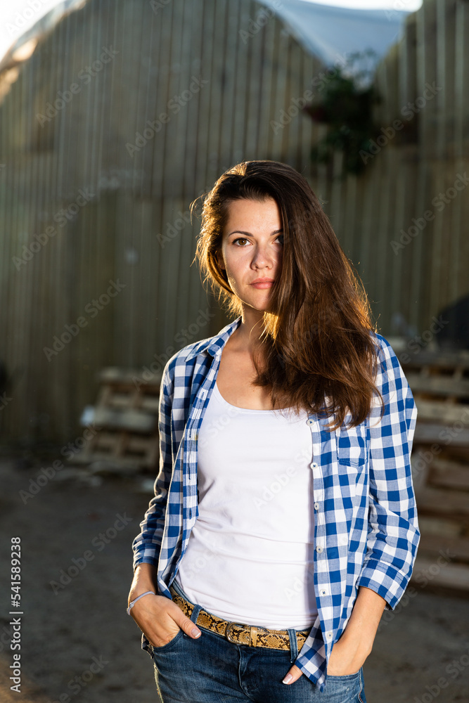 Attractive brunette wearing blue checkered shirt posing in farm against background of greenhouses