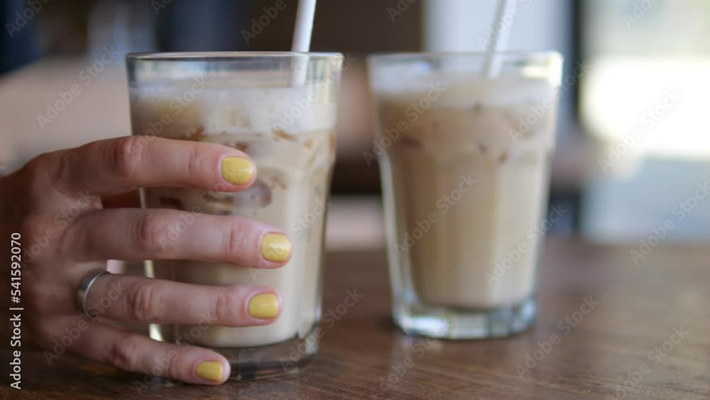 On a wooden table are two full glasses with a plant-based milkshake and ice cubes. A woman's hand takes one of the glasses and moves it towards herself. On a wooden table are two full glasses with a plant-based milkshake and ice cubes. A woman's hand takes one of the glasses and moves it towards herself.