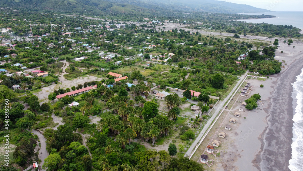 Panoramic view of green, tree covered landscape, houses, buildings, roads and black sandy beach, aerial drone views of Liquica town in Timor Leste, Southeast Asia