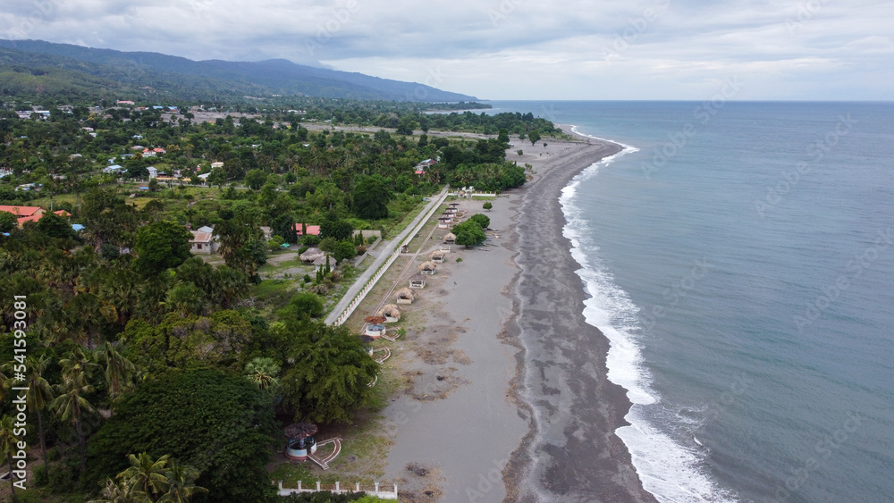 Aerial drone landscape view of black sandy beach, ocean waves and tree covered terrain in town of Liquica, Timor Leste, Southeast Asia