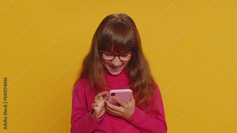Young preteen child girl kid use mobile smartphone typing browsing say Wow yes found out great big win good news celebrate doing winner gesture. Little toddler children on studio yellow background Young preteen child girl kid use mobile smartphone typing browsing say Wow yes found out great big win good news celebrate doing winner gesture. Little toddler children on studio yellow background