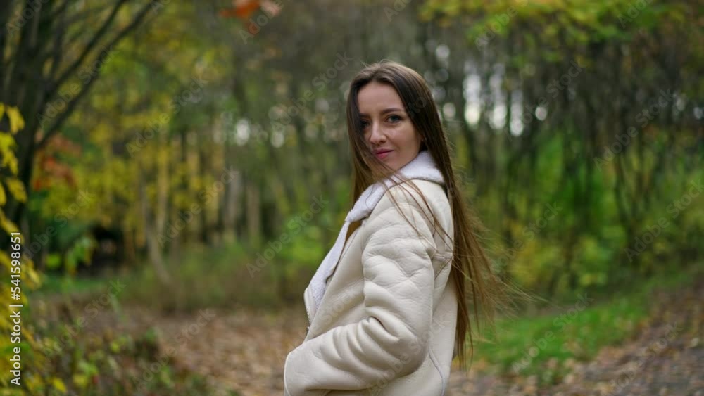 Long-haired brunette woman standing in the autumn park turns around to the camera. Lady looks into camera slowly approaching to her face. Blurred backdrop. Long-haired brunette woman standing in the autumn park turns around to the camera. Lady looks into camera slowly approaching to her face. Blurred backdrop.